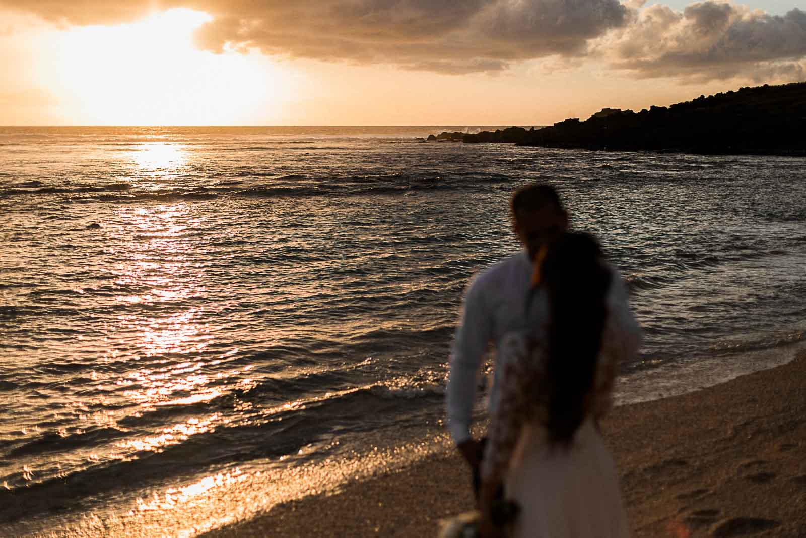 Photographie de Mathieu Dété, photographe de mariage sur l'île de la Réunion 974, présentant un couple de marié au premier plan, flou, et la vue sur le coucher de soleil en arrière plan, à Saint-Leu