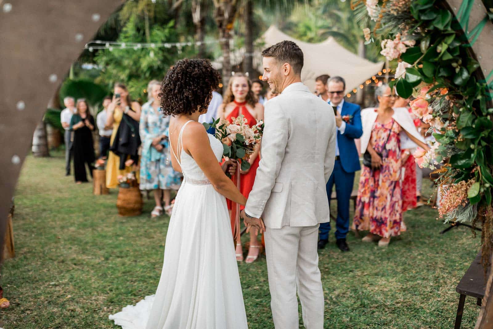 Photographie de Mathieu Dété, photographe de mariage et grossesse à Sainte-Marie, sur l'île de la Réunion 974, présentant les mariés main dans la main sous l'arche de la cérémonie laïque, au Jardin d'Ama à Saint-Leu, devant leurs invités