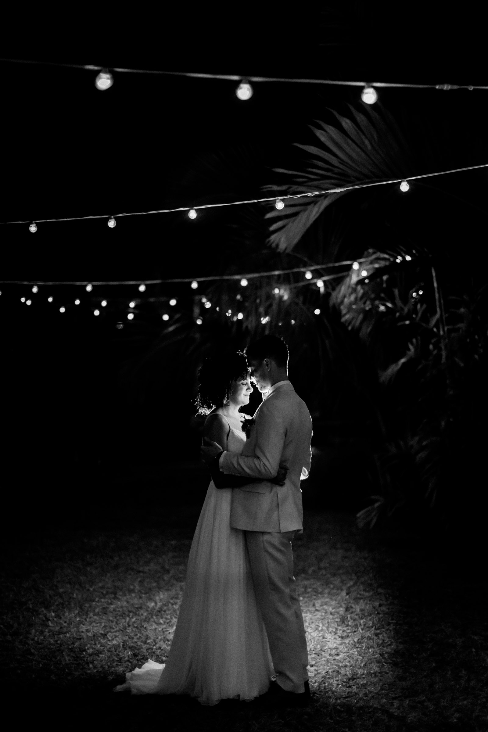 Photographie de Mathieu Dété, photographe de mariage à Saint-Pierre sur l'île de la Réunion 974, présentant un couple de marié en noir et blanc sous les guirlandes lumineuses du Jardin d'Ama, à Saint-Leu