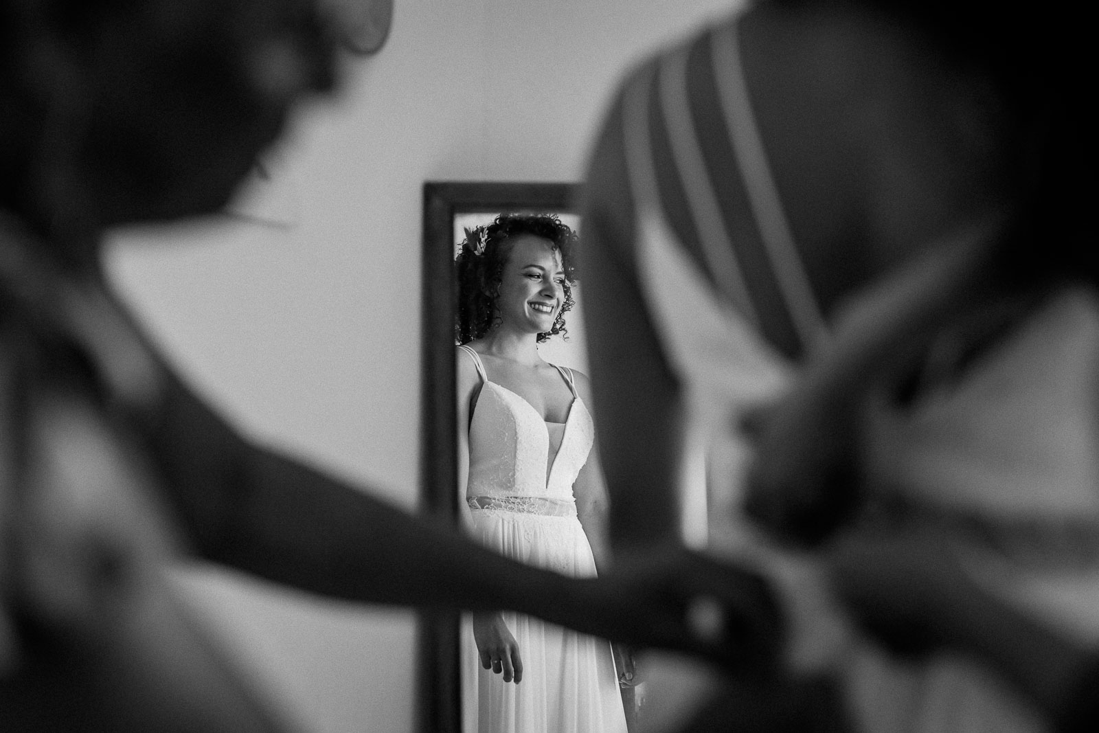 Photographie de Mathieu Dété, photographe de mariage et grossesse à Sainte-Marie, sur l'île de la Réunion 974, présentant la mariée qui sourit dans le miroir en se faisant accrocher la robe, en noir et blanc