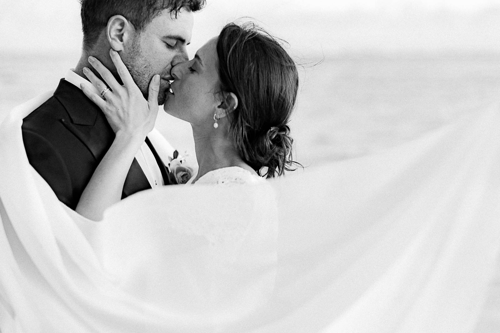 Photographie en noir et blanc de Mathieu Dété, photographe de mariage à Saint-Leu sur l'île de la Réunion 974, présentant un couple de mariés qui s'embrasse, avec le voile de la mariée au premier plan