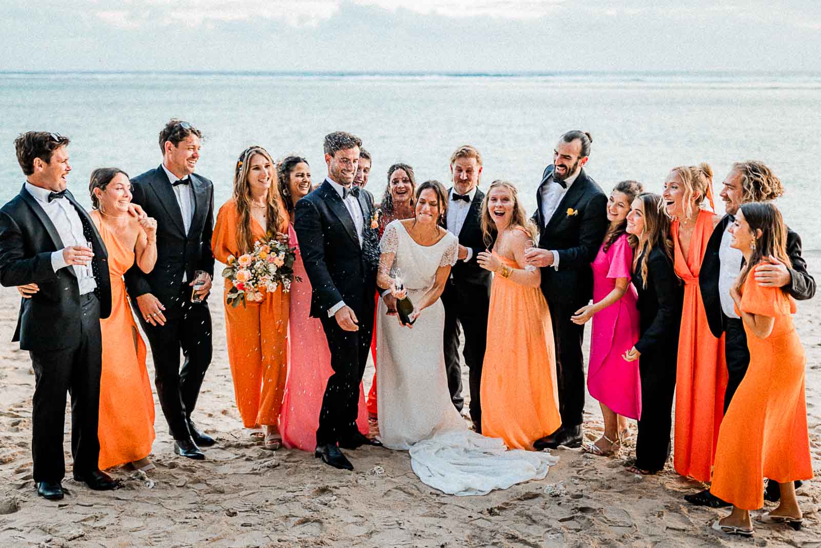 Photographie de Mathieu Dété, photographe de mariage à Saint-Leu sur l'île de la Réunion 974, présentant une mariée qui ouvre une bouteille de champagne, entourée de ses invités, sur la plage du LUX* Saint-Gilles