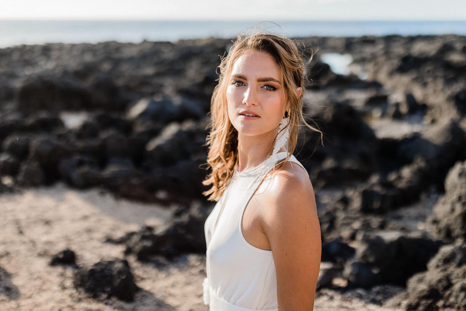 Photographie de Mathieu Dété, photographe de mariage, de mode et de fiançailles à Saint-Paul sur l'île de la Réunion 974, présentant Emma, mannequin, en robe de mariée de la marque Maison Mananne, se retournant face à l'objectif, au milieu des roches de la cote de la Réunion